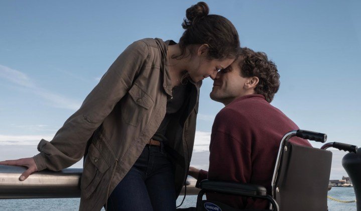 Una mujer apoya su cabeza contra la cabeza de un hombre que está sentado en una silla de ruedas.