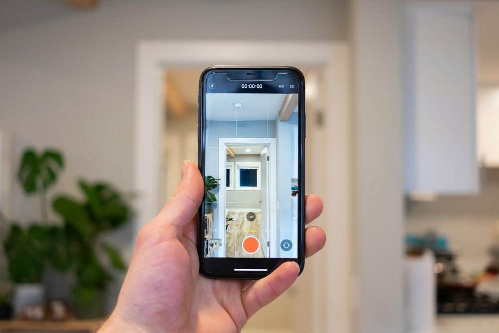 Mano de una persona sosteniendo un smartphone que muestra el panel de control de una aplicación de casa inteligente, con la sala de estar de fondo.