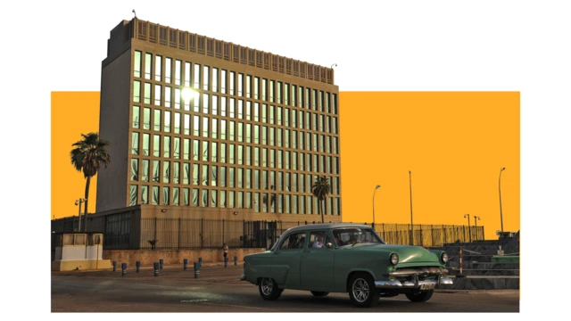 Fotografía de la Sección de Intereses de Estados Unidos en La Habana, tomada el 1 de julio de 2015. Un coche verde antiguo pasa junto al edificio de hormigón, que cuenta con altas ventanas de cristal y está rodeado de algunas palmeras.