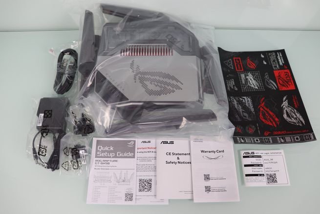 Contenido completo de la caja del router ASUS GT-BN98 sobre una mesa blanca, incluyendo cables, fuente de alimentación y manuales.